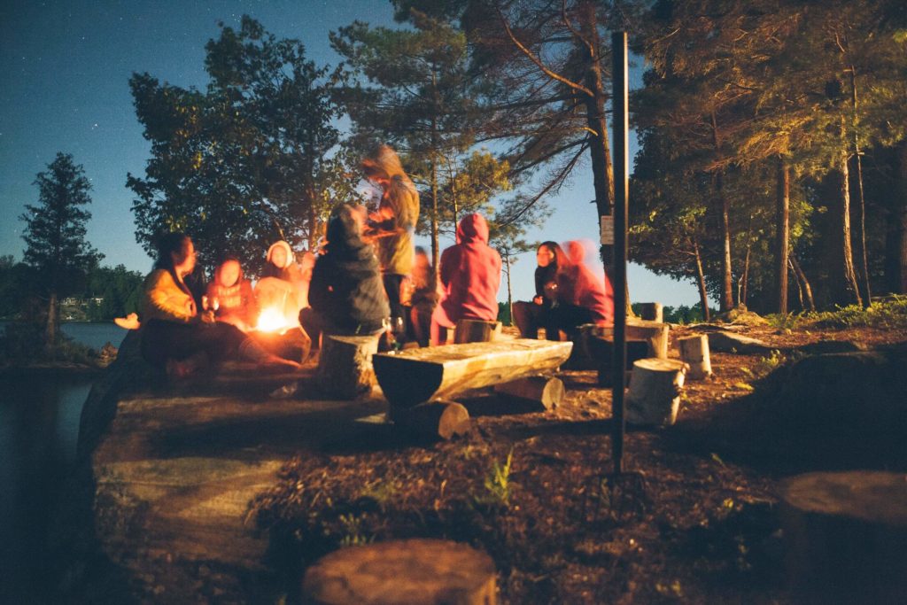 eine Gruppe von Menschen sitzt in der Abenddämmerung im Wald um ein Lagerfeuer und unterhält sich
