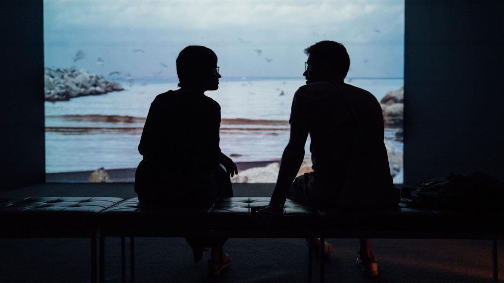two people with glasses sit on a bench in front of a screen 