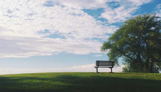 a bench on green grass overlooking from the top of a hill, with clouds and blue sky