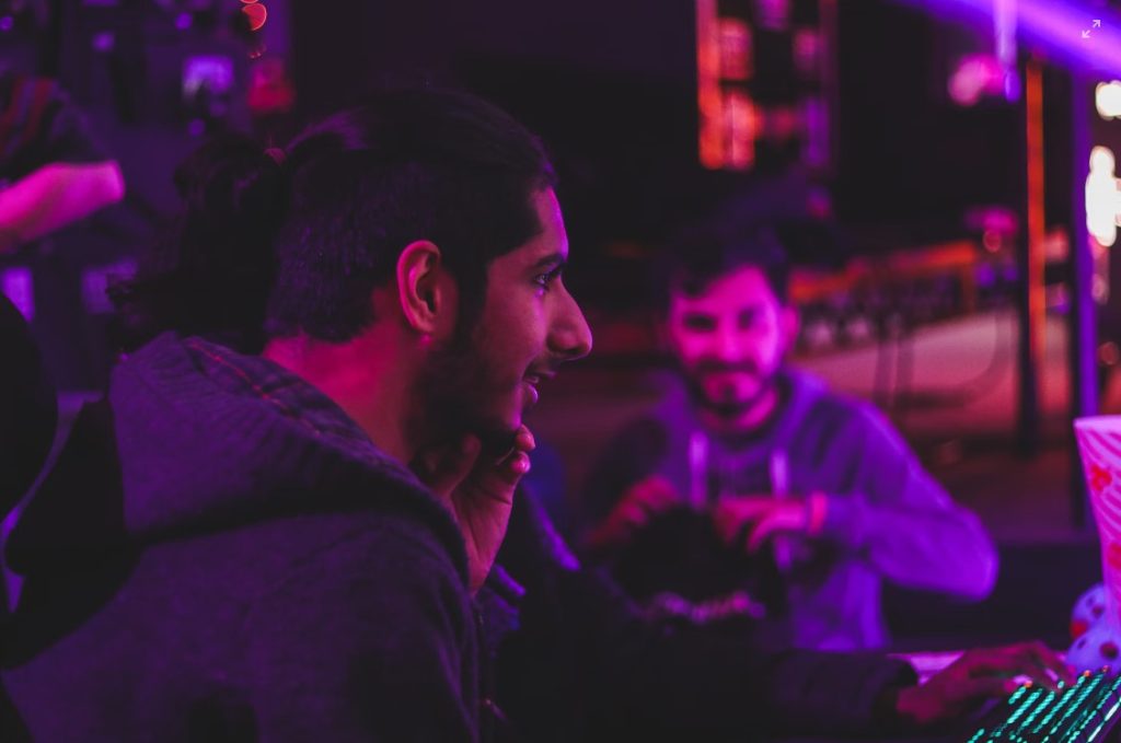 une personne souriante regardant un écran d'ordinateur avec des gens en arrière-plan. Une lumière rose projette une ombre froide sur eux.