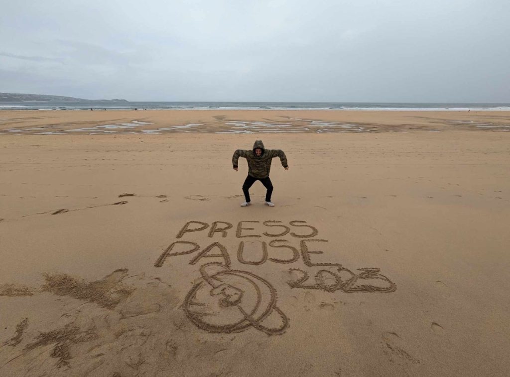 someone is stood on a beach, with Press Pause 2023 and a SIOW planet drawn in the sand. 