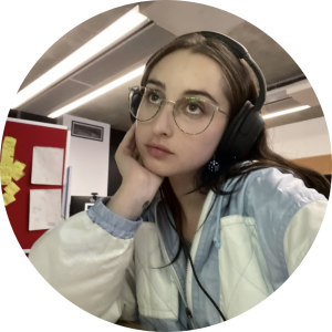 A photo of Julia wearing a white and blue jacket and black headphones, resting their face in their hand and looking up.